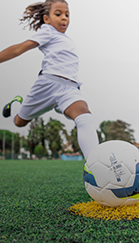 Marca exclusiva Decathlon: Kipsta. Criança com uniforme branco de treino chutando bola de futebol no campo gramado.