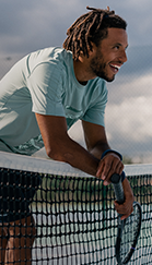 Marca exclusiva Decathlon: Artengo. Homem negro com dreads sorrindo veste camiseta na cor azul clara apoiado na rede de tênis com sua raquete em mãos.