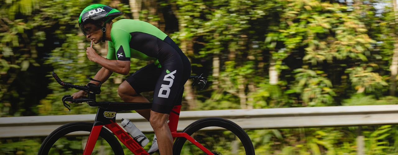 Só na Decathlon online: Homem branco uniformizado com o patrocionio da Dux Nutrition realizando passeio de bicicleta e consumindo um gel de carboidrato para ganho de performance